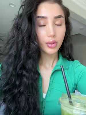 A woman with long, dark, wavy hair and closed eyes is the main subject. She's wearing a green zip-up top. A beverage in a clear glass with a straw is visible.