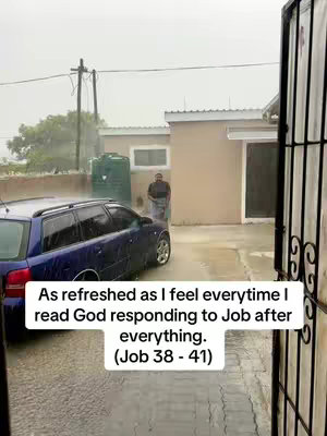 An outdoor scene shows a dark blue station wagon parked on a wet surface next to a two-story building. A man, appearing to be the video creator, is standing further in the background, and it appears like it is raining. Text overlayed on the image expresses a sentiment related to scripture, specifically, "As refreshed as I feel everytime I read God responding to Job after everything. (Job 38 - 41)"