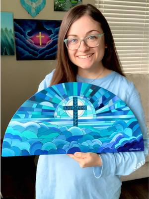 A woman with brown hair and glasses is holding a light-blue, semi-circular painting with a cross and sun in front of her. She is smiling. Behind her appears to be more artwork on beige walls.  She wears a light-blue long-sleeve top. 