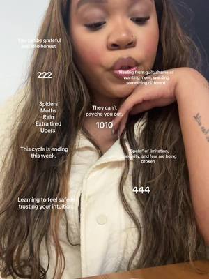 A selfie of a woman with long, wavy brown hair. She is holding her phone and looking toward the camera with her mouth slightly open. Text overlays are scattered across the image, including numbers and phrases related to spirituality and self-reflection. The background appears to be a neutral-colored wall.