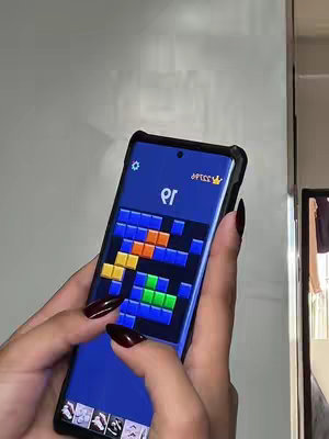 A close-up shot showcasing a hand, with polished maroon nails, holding a smartphone displaying a mobile game. The game screen shows colorful blocks within a grid, typical of a puzzle or matching game. The background is a plain white surface. The phone is enclosed in a black, protective case, and the overall composition is simple and focused.