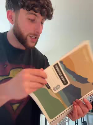 A young man with curly hair and a short beard is holding a notebook and a card. He's looking at the camera and talking. The notebook has a patterned cover. The background is a plain wall.