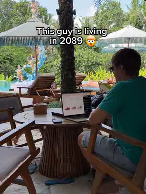 A man is sitting at a table with a laptop, working outdoors. The background shows a pool, umbrellas, and trees. The hook text 'This guy is living in 2089' is overlaid on the scene.