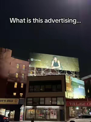 An outdoor shot at night featuring buildings and a large billboard. The billboard displays an advertisement for Cash App, featuring a person. Above is a text overlay 'What is this advertising...'