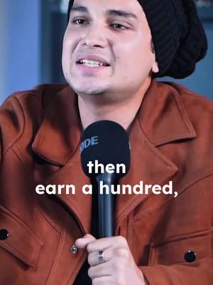 A man in a brown jacket and a black beanie is holding a microphone in front of him. There's a text overlay on the bottom of the image.