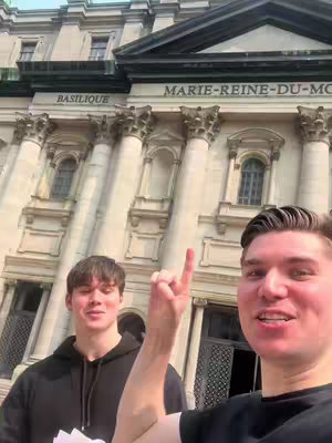Two young men stand in front of a large building with columns. One man is pointing upwards. The building has ornate details and the words "BASILIQUE MARIE-REINE-DU-MONDE" are visible.