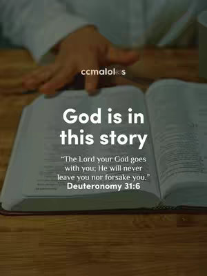 An open Bible on a wooden table. The left page of the Bible is visible, and the text 'God is in this story' is overlaid, along with a Bible verse from Deuteronomy. A hand is visible on the top, near the pages.