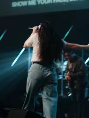 The cover image shows a woman on stage performing with a microphone. She is seen from the back with her long, dark curly hair. She's wearing a light-colored top and jeans. The stage has lights and what looks like a screen with text on it. Other band members can be slightly seen in the background.