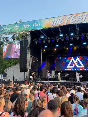 The image captures a concert or festival atmosphere, with a large stage as the central focus. The stage is brightly lit, with LED screens displaying visuals and the event's branding, and a crowd of people fills the foreground, suggesting an energetic live performance.