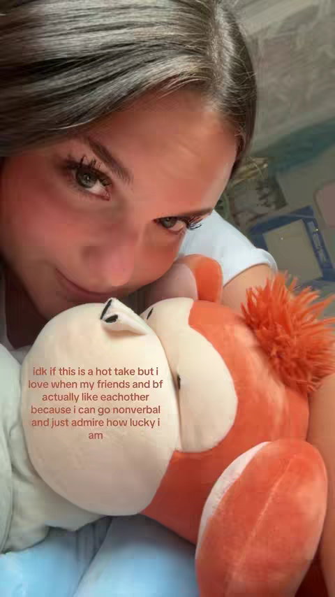 A young woman is shown in a close-up, embracing a plush toy with an orange and white color scheme. Her face fills most of the frame, with her eyes looking towards the viewer. Text overlayed on the bottom half of the image reads, “idk if this is a hot take but i love when my friends and bf actually like eachother because i can go nonverbal and just admire how lucky i am.” The overall composition is warm and intimate.