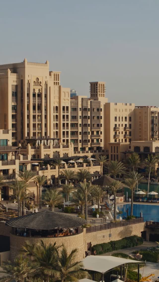An aerial view of a luxury resort in a desert location. The architecture is primarily beige with intricate Arabesque designs. Palm trees are scattered throughout the property, and several pools and outdoor seating areas can be seen. The sky is a clear, pale blue.