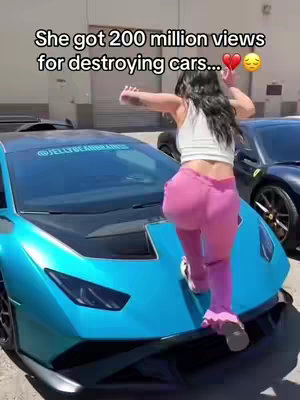 A woman is shown standing on the hood of a teal sports car, leaning towards the right side of the frame. She is wearing a white crop top and pink pants. The background includes other cars and a building.