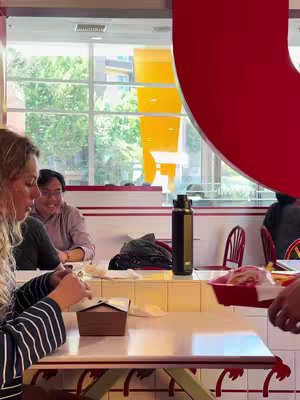 The image features a blurred foreground with a person seemingly eating at a table inside a restaurant. Two other people are seated, smiling and facing the same direction as the person in the foreground. The restaurant has a red and white theme, with large windows showing a glimpse of the outside, and with a big red shape on one side.