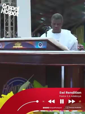 The image shows a man in a white outfit standing behind a wooden podium. The man is looking down, appearing to be reading from something. The background is slightly blurred, with hints of a church setting. A red bar at the bottom features video controls and text identifying the video as 'Ewi Rendition' by 'Pastor E.A Adeboye'. A logo for 'Gospel Hotspot' is in the top left.