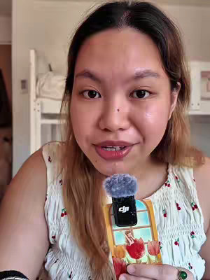 A woman with light skin and long, light-brown hair is holding a microphone to her face. She is wearing a white tank top with red cherry patterns. The woman is looking at the camera with a slightly open mouth as if speaking. A microphone, with a fluffy grey cover, is partially visible. Behind the woman is a blurry white bunk bed.