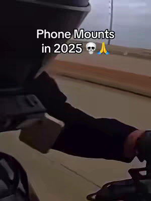 The image shows a partial view of a person's arm and hand inside a vehicle, possibly driving. Text overlayed on the image reads "Phone Mounts in 2025", followed by a skull and praying hands emoji. The background is the interior of the vehicle and possibly an abstracted view of the road.
