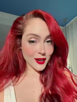 A close-up shot featuring a fair-skinned woman with vibrant red hair, striking makeup, and a slight smile. Her hair is styled in loose waves, framing her face. She wears bold red lipstick, prominent winged eyeliner, and has a slight blush, all creating a polished appearance. The background is blurred, suggesting a shallow depth of field.