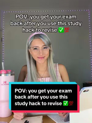 A young woman with fair skin, glasses, and silver-toned hair sits at a desk, smiling at the camera. The background is simple, with a wall lit by a string of lights. In front of her is a pink Stanley tumbler, and a tablet on the desk. Her expression is friendly and approachable.