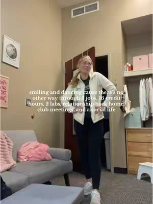 A young woman is standing in a dorm room, smiling and looking up. She is wearing a white jacket, black pants, and sneakers. The room has light-colored walls, a gray couch, and a door in the background. Text overlays the image.