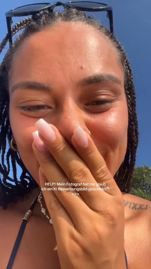A selfie-style shot featuring a young woman covering her mouth with her hand. She has medium-length, braided hair with sunglasses perched on her head. The background is a bright blue sky, suggesting a sunny day.