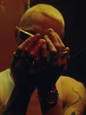 A close-up shot of a person whose face is partially obscured by their hands, which are covered in a red substance, possibly blood. The person is wearing sunglasses, and the lighting is dim, emphasizing the contrast.