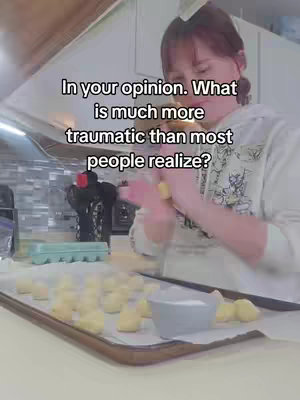A woman in a kitchen scene is the focus. She is wearing a white hoodie and working with dough. The background shows kitchen cabinets, a counter, and a window providing natural light.