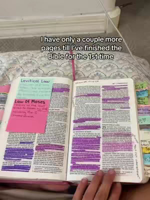 The image shows an open Bible on a bed with highlighted passages and sticky notes. The Bible is open to a section with text highlighted in purple. Several colored sticky notes are attached to the pages, and a pink bookmark is inserted. A hand is visible, holding the book open. The background shows a white surface that might be a bed headboard.