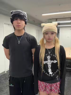 Two young people stand side-by-side, facing the camera in a brightly lit interior setting. The individual on the left is male, wearing a black t-shirt and black pants. The individual on the right is female, wearing a black long-sleeved shirt, mini skirt, and light-colored beanie hat. Both have a serious expression. Each of them is wearing a cross necklace. The background is clean, with an open door visible in the hallway.