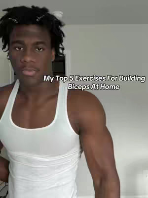 The image shows a young Black man in a white tank top. The man is looking at the camera. The text overlay says "My Top 5 Exercises For Building Biceps At Home"