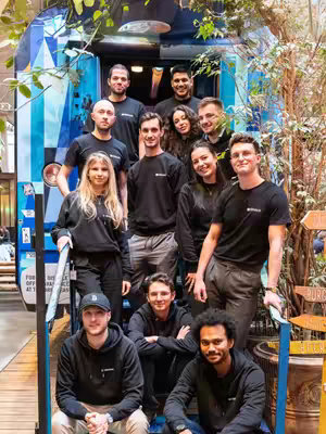 A group of people, presumably employees, posing on a staircase in what appears to be a casual office setting. They are of varying ages and genders, dressed in a mix of black t-shirts and hoodies. The background includes architectural details and partial views of a building exterior. The composition uses a medium shot to capture the whole staff. The overall lighting is natural, and the color palette is neutral with a few pops of nature green.