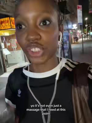 A selfie shot of a person speaking on a city street at night, wearing a black and white Adidas shirt and earbuds.