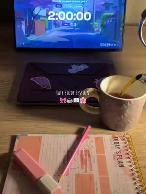 A top down view of a workspace on a wooden surface. In the foreground is a pink planner with the words "Today's Plan" visible. A pink pen and highlighter rest on top of it. To the right is a cup with something being poured into it. Behind the cup is a laptop. On the laptop screen is a digital timer which reads 2:00:00. The background is a representation of a street in a town.