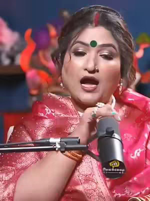 A woman with dark hair, adorned with a red bindi and a green dot on her forehead, is holding a microphone. She's wearing a red blouse and gold jewelry, with an open mouth, as if singing or speaking. The background is blurred, suggesting a studio setting with colorful decorations.