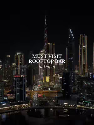 An eye-level shot of Dubai's skyline at night, featuring illuminated skyscrapers against a dark sky. Overlayed text reads "MUST VISIT ROOFTOP BAR in Dubai". General ambiance is dark, emphasizing the city lights.