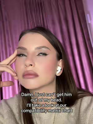 A medium shot features a young woman with brown hair and fair skin. She has makeup on with dark eyeliner and a light lip color. The background is a pink curtain. She is wearing a beige sweater and has a white AirPod in her right ear. She is looking down and left, with her right hand touching her temple and some text overlaid at the bottom.