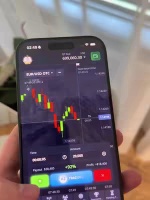 A person's hand holding a smartphone displaying a trading app's interface. The screen shows a candlestick chart with green and red bars, along with financial data such as price fluctuations, time, and potential profit figures. The environment looks like a home setting.