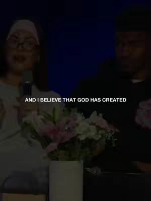 The cover image shows two people, a woman and a man, partially visible, engaged in a conversation. The woman on the left, is wearing glasses and a head covering, with a microphone in front of her. A bouquet of flowers sits in the middle. Text overlayed at the bottom reads, 'AND I BELIEVE THAT GOD HAS CREATED'. The background is dark, suggesting a low-lit indoor setting.