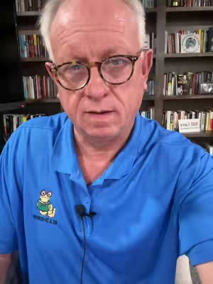 A man wearing glasses and a blue polo shirt stands in front of a bookcase. He is looking at the camera. There is a microphone clipped to his shirt.