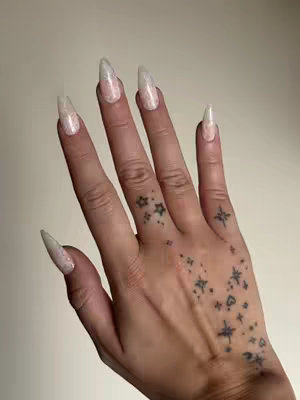 A close-up shot of a hand with long, glittery acrylic nails. The hand is angled slightly, and the fingers are spread. The nails have a light, shimmering finish. On the back of the hand are small star tattoos. The background is a plain, neutral color.