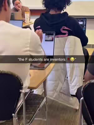 A classroom setting with students at desks. One student is in the foreground with a laptop, with the back of the person in a black hoodie with a pink design visible. The hook shows up at the bottom. Additional student is visible in the background, also using a laptop. The overall tone is casual, and the lighting appears to be from the classroom.
