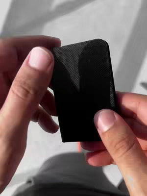 A person's hands hold a small, rectangular black object, possibly a wallet or a tech device. It appears to be a Ledger device; the light is reflecting off of the material. The background is a neutral white surface, which provides a clear view of the hands and the object.