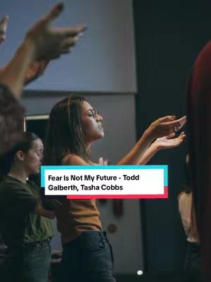 A woman with glasses stands with her arms raised, eyes closed, in what appears to be a worship setting. Other figures are blurred in the background with raised hands, and overlaid text identifies the song and artists.