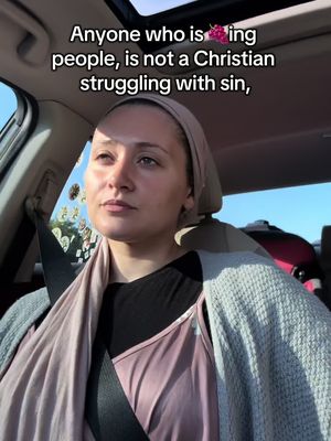 A woman in a car looks pensive. She wears a headscarf and a light grey sweater. The background shows a sunny day and surrounding trees.