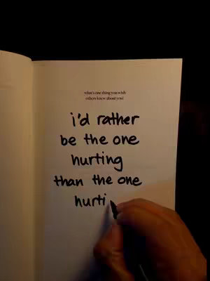 An open notebook page with handwritten text in black ink. The text reads, 'i'd rather be the one hurting than the one hurting'. The hand of a person is visible at the right of the paper, holding a pen. The background is dark and out of focus.