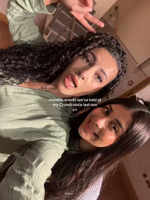 Two young women are seen smiling at the camera, positioned closely together against the muted backdrop of a room, likely indoors.