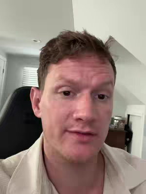A medium shot shows a man, centered in the frame, looking directly at the viewer. He has close-cropped brown hair and is wearing a light-colored, collared shirt. The background is blurred.