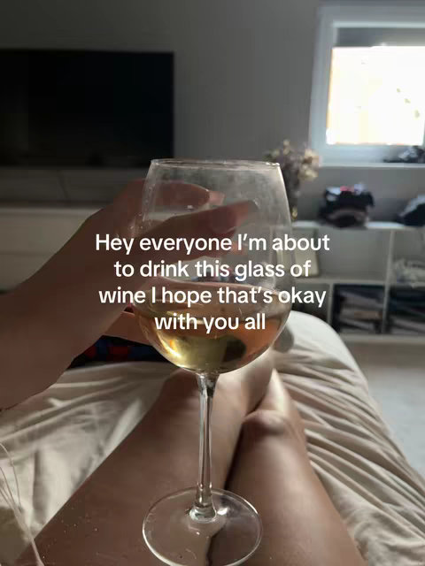 A person is lying on a bed. They are holding a wine glass, filled with a light-colored liquid, possibly white wine, in their right hand. The person's legs are visible, and the bed appears to be covered in a light-colored sheet. The background is slightly blurred, suggesting an indoor setting.