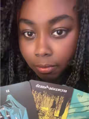 The cover image features a close-up of a person's face, focusing on their eyes and the tarot cards they are holding. The person has dark skin, dark eyes, and long braided hair. They are looking down towards the cards, which are held in front of them. The overall color tone is warm with natural lighting.