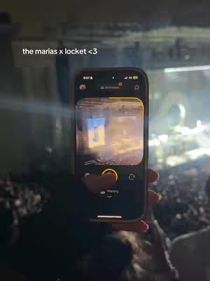 A person holding a phone up at a concert. The phone screen shows a photo from locket. The background behind the phone shows the crowd and stage during a concert.