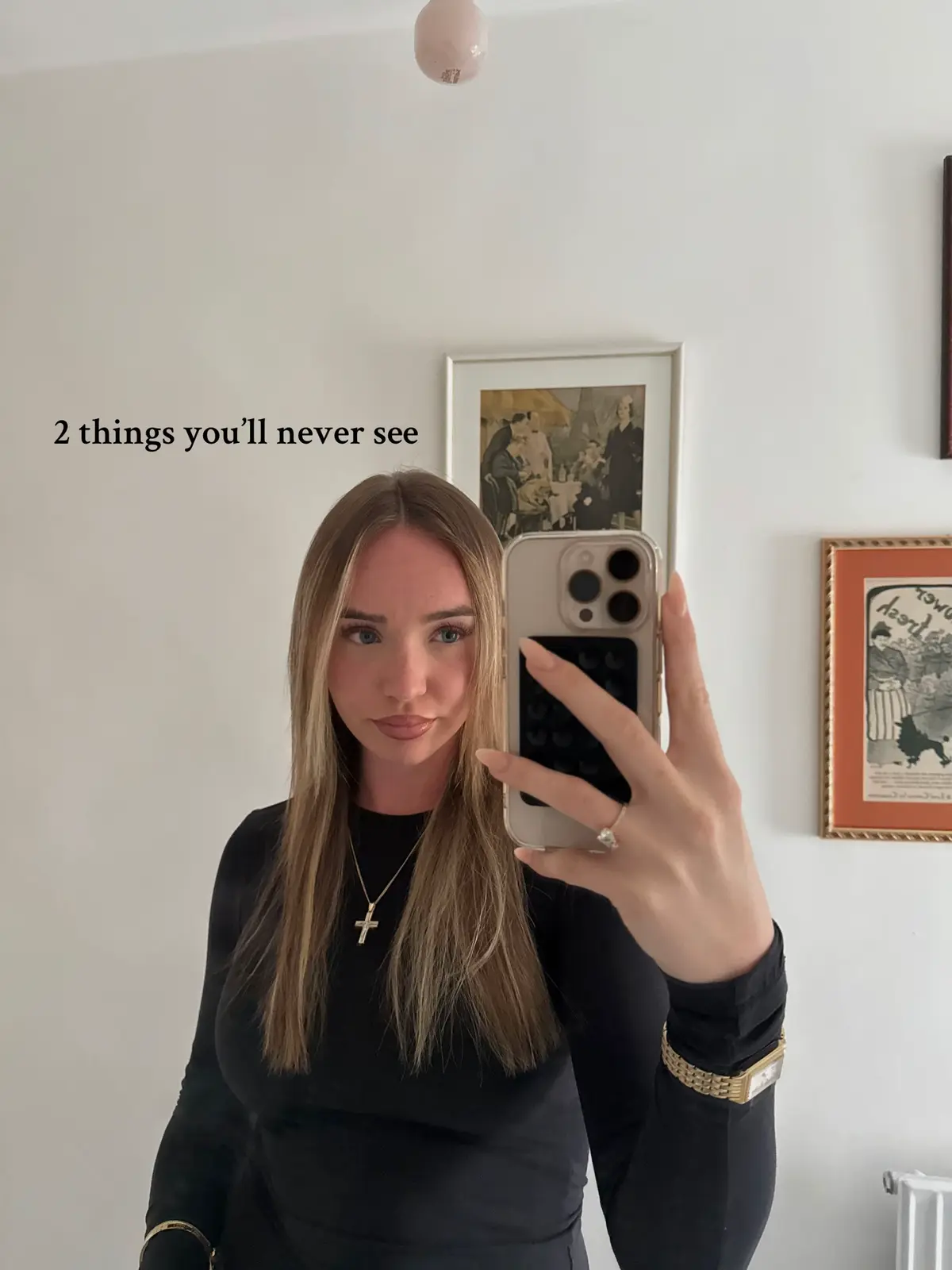 A woman is taking a selfie in front of a mirror. Her hair is long and blonde with slight waves. She wears a black top and a gold necklace with a cross pendant. She is holding a phone in front of her face. There is text overlayed at the top left of the image. In the background, there are framed pictures on the white wall. One of the frames contains a black and white picture. 